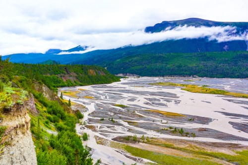 A winding river flows through a lush green valley, surrounded by mountains and cloudy skies.
