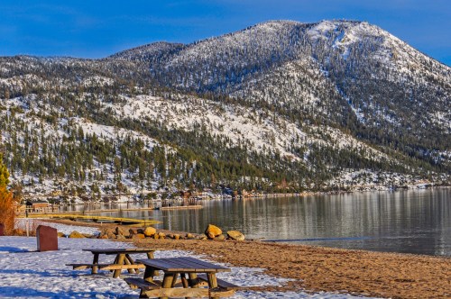 A serene lakeside scene with snowy mountains, a sandy beach, and picnic tables under a clear blue sky.