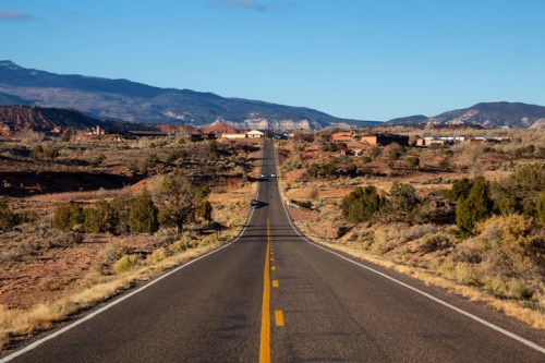 A long, straight road stretches through a desert landscape with mountains in the background under a clear blue sky.