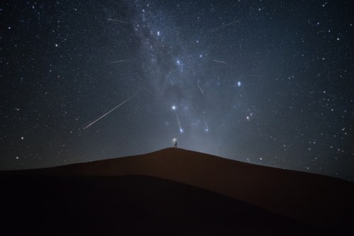 A silhouette of a person on a sand dune under a starry sky with shooting stars and the Milky Way visible.