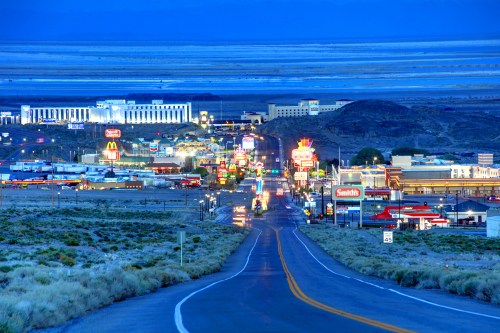 A winding road leads to a brightly lit town at dusk, surrounded by mountains and open land.