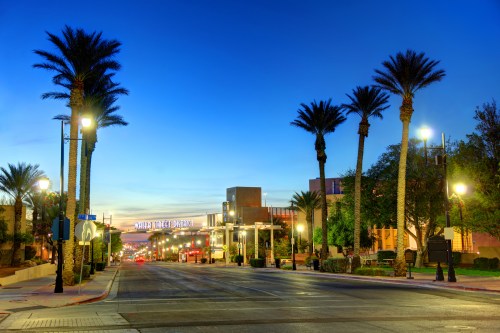 A quiet street lined with palm trees, illuminated by streetlights, at dusk with a colorful sky in the background.