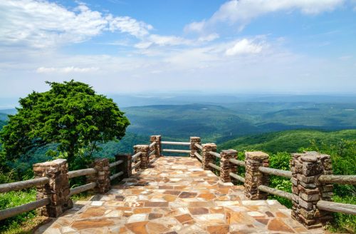 Scenic overlook with stone railing, lush green mountains, and a blue sky dotted with clouds in the background.