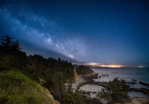 A starry night sky over a coastal landscape, with the Milky Way visible and waves gently lapping at the shore.