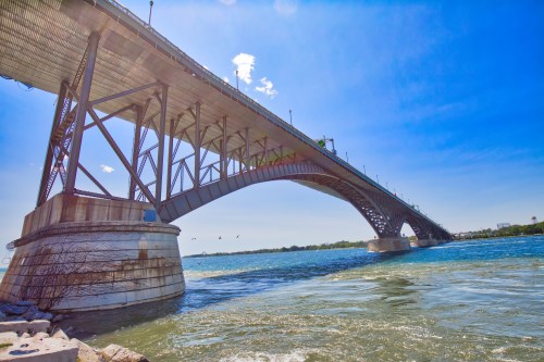 A large bridge arches over a river under a clear blue sky, with water and a few birds visible below.