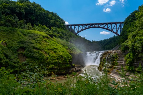A scenic view of a waterfall surrounded by lush greenery, with a bridge spanning above under a clear blue sky.