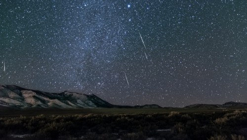 A starry night sky filled with bright stars and several shooting stars over a mountainous landscape.