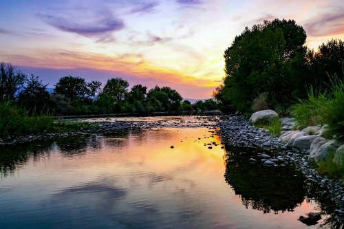 A serene river at sunset, reflecting colorful skies and surrounded by trees and rocky banks.
