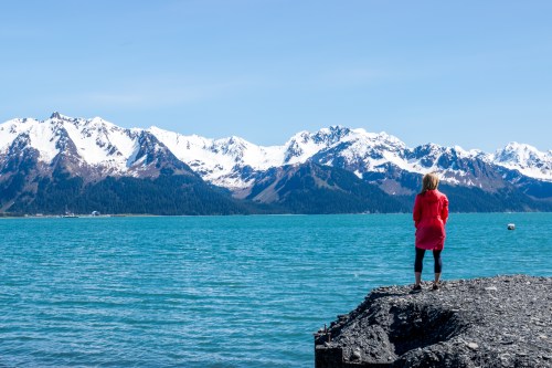 A person in a red jacket stands on a rocky shore, gazing at snow-capped mountains across a turquoise sea.