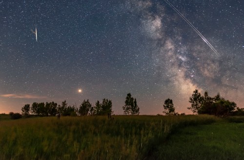 A starry night sky with the Milky Way, a comet, and satellite trails over a grassy field and trees.