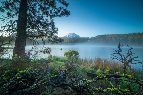 A serene lake scene at dawn, with mist rising, mountains in the background, and lush greenery in the foreground.