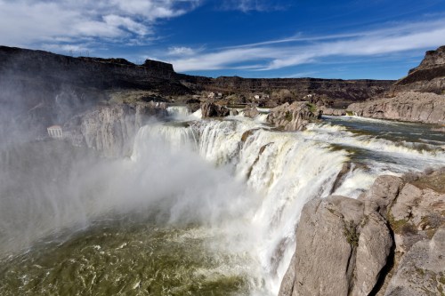 A stunning waterfall cascades over rocky cliffs, surrounded by a scenic landscape and blue sky with wispy clouds.