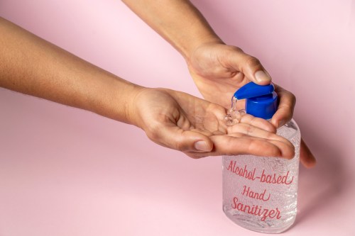 A person pours hand sanitizer from a bottle into their palm against a pink background.