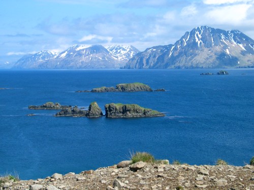 A scenic view of rocky islands in a blue ocean, surrounded by snow-capped mountains under a partly cloudy sky.
