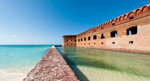 Historic brick fortification by the sea, with clear turquoise water and a blue sky in the background.