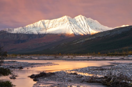 Snow-capped mountains reflect golden light at sunset, with a serene river and rocky foreground.