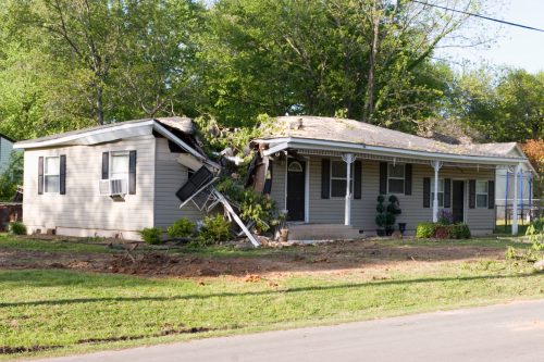 A house with a damaged roof and fallen tree branches, surrounded by a grassy yard and trees.