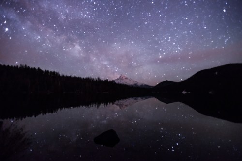 A starry night sky reflects on a calm lake, with a snow-capped mountain in the background.