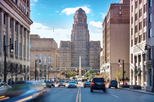 A busy city street with cars, leading to a tall, historic building under a clear blue sky.