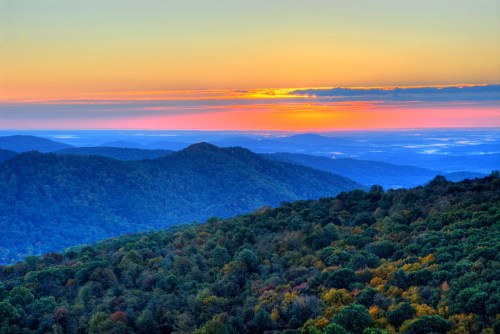 A vibrant sunset over rolling mountains, with colorful foliage in the foreground and a serene sky.