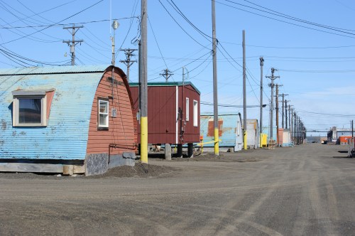 Row of colorful, curved-roof buildings along a dirt road, with power lines stretching overhead under a clear blue sky.