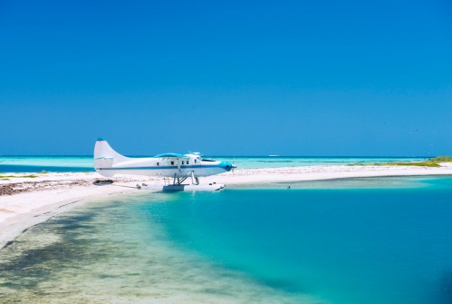A seaplane parked on a sandy beach beside clear turquoise waters under a bright blue sky.