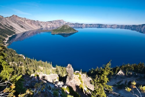 A panoramic view of Crater Lake, featuring deep blue water and a small island surrounded by rugged cliffs and trees.