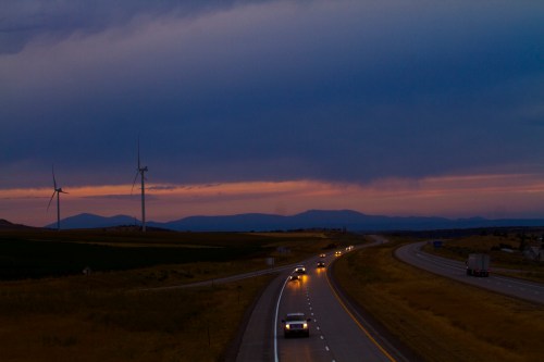 A highway at dusk with cars driving, wind turbines in the background, and mountains under a cloudy sky.