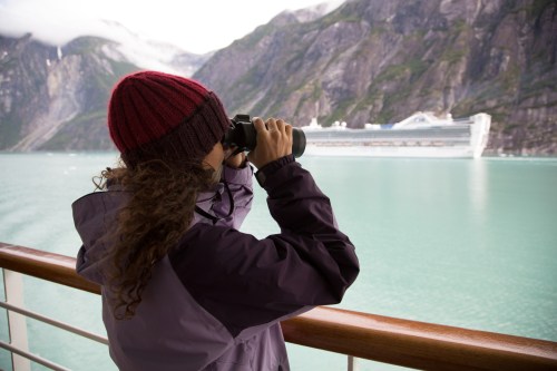 A person in a maroon beanie uses binoculars to observe a cruise ship in a scenic, mountainous landscape.