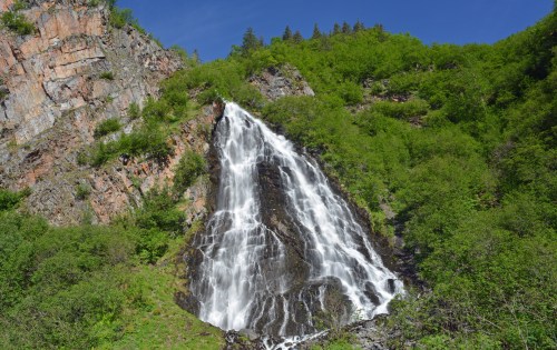 A cascading waterfall flows down a rocky cliff, surrounded by lush green foliage and a clear blue sky.