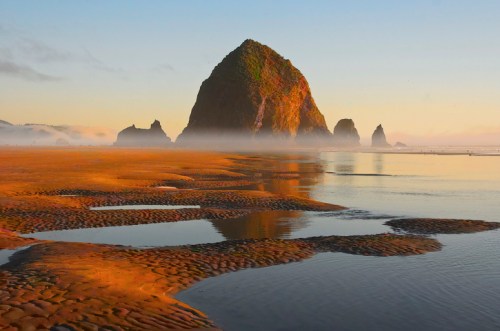 A serene beach scene at sunset, featuring a large rock formation and calm waters reflecting the colorful sky.