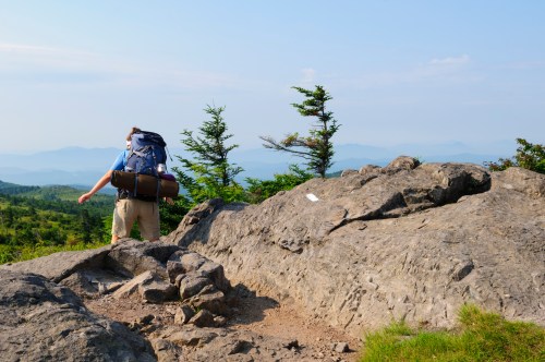 A hiker with a backpack stands on a rocky outcrop, overlooking a scenic mountain landscape under a clear blue sky.
