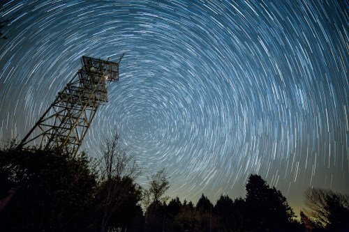 A starry night sky with circular star trails above a tall radio tower and silhouetted trees.
