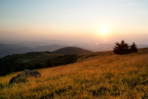 A serene landscape at sunset, featuring rolling hills, a grassy field, and distant mountains under a colorful sky.