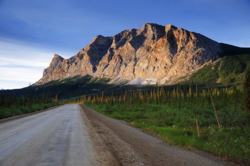 A dirt road leads towards a towering mountain, illuminated by sunlight, surrounded by lush greenery and blue sky.