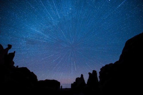 Starry night sky with streaks of light radiating from a central point, framed by dark rock formations.