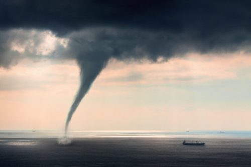 A waterspout forms over the ocean, with a ship visible in the distance under a cloudy sky.