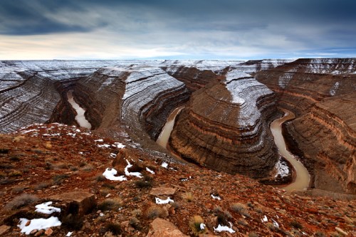 A dramatic canyon landscape with winding rivers, layered rock formations, and patches of snow under a cloudy sky.