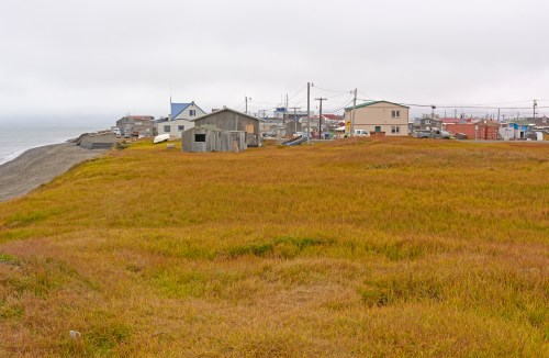 A coastal village with colorful houses, grassy fields, and a cloudy sky overlooking the ocean.