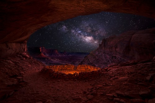 A stunning night sky filled with stars and the Milky Way, viewed from a rocky cave overlooking a desert landscape.