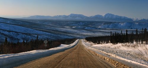 A winding dirt road leads through a snowy landscape, with mountains in the distance under a clear blue sky.