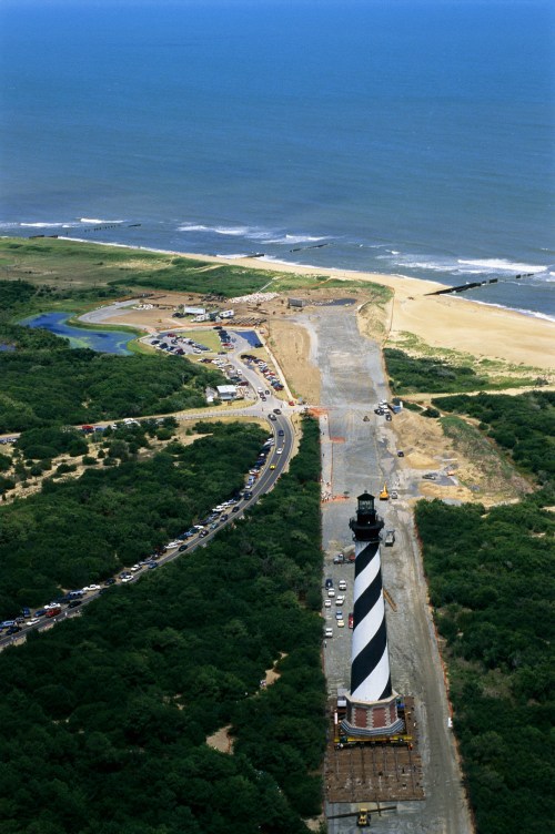 Aerial view of a striped lighthouse near the coast, surrounded by greenery and a sandy beach.