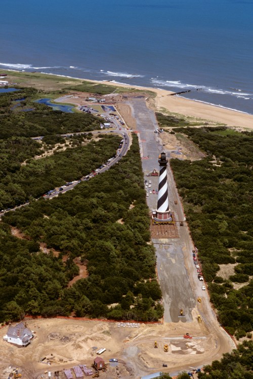 Aerial view of a lighthouse near the coast, surrounded by trees and a road, with the ocean in the background.