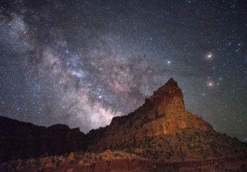 A starry night sky filled with the Milky Way over a rugged rock formation in a dark landscape.