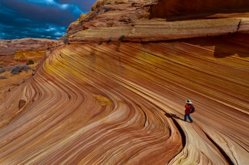A person in a hat walks along colorful, swirling rock formations under a dramatic sky.