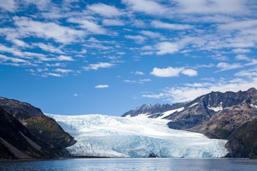 A vast glacier flows down from mountains under a blue sky with scattered clouds, reflecting in calm waters below.