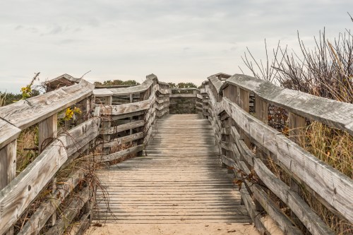 A wooden boardwalk leads through tall grass to a sandy beach under a cloudy sky.