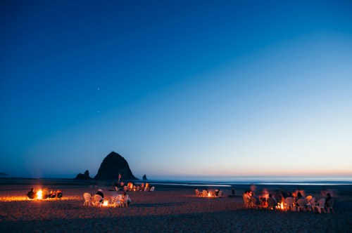 A beach at dusk with people gathered around fires, silhouetted against a rocky outcrop and a clear blue sky.