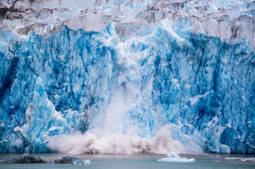 A massive glacier calving into the water, creating a splash and revealing vibrant blue ice formations.