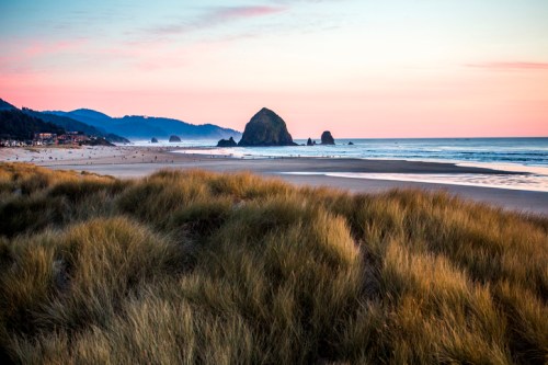 A serene beach scene at sunset, featuring gentle waves, grassy dunes, and a prominent rock formation in the distance.